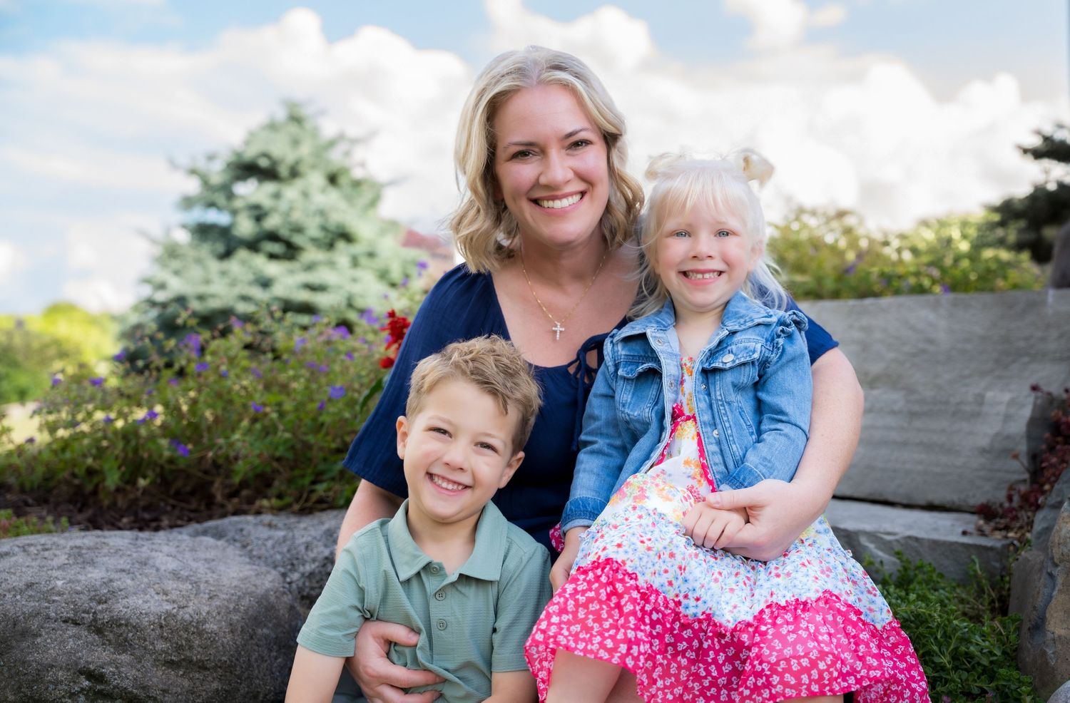 A smiling woman poses outdoors with two young children, a boy and a girl, in a lush garden setting.