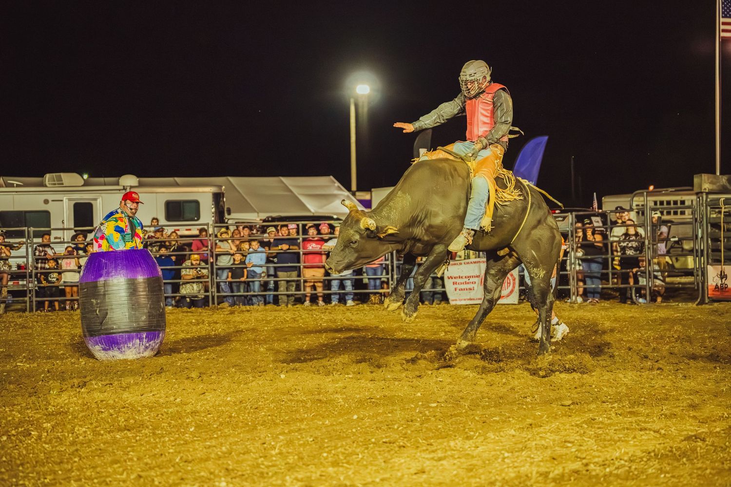 A bull rider competes at a nighttime rodeo, gripping a bucking bull near a colorful barrel clown.