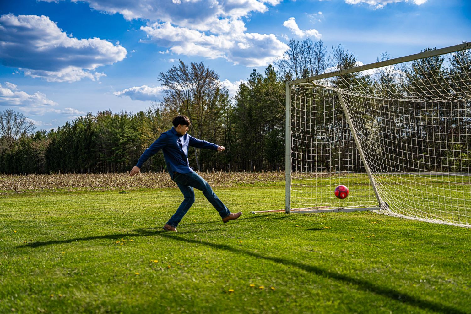 Young man in blue suit kicking a red soccer ball toward a goal net on a green field.