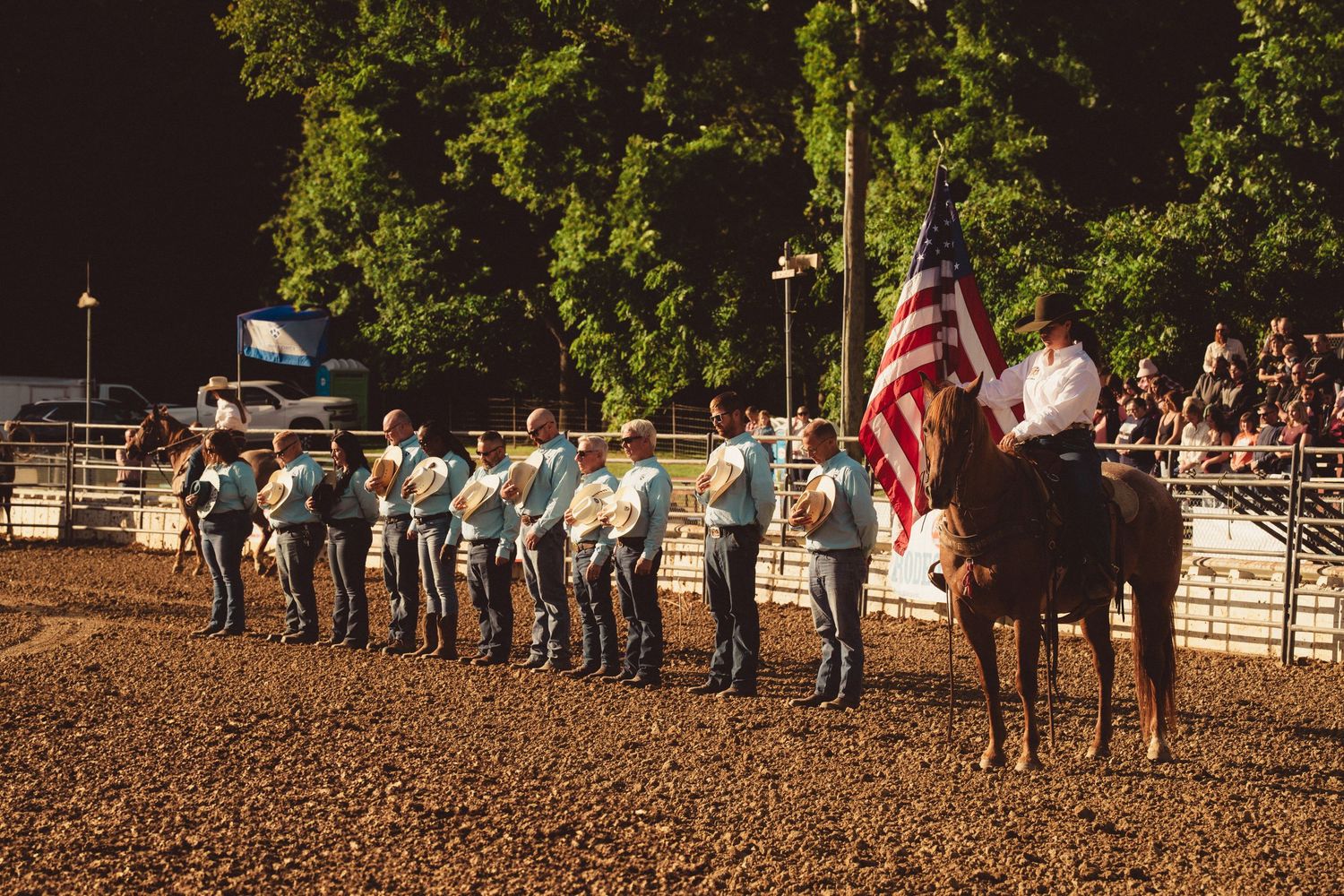 Cowboys line up with an American flag on horseback at a rodeo opening ceremony at dusk.