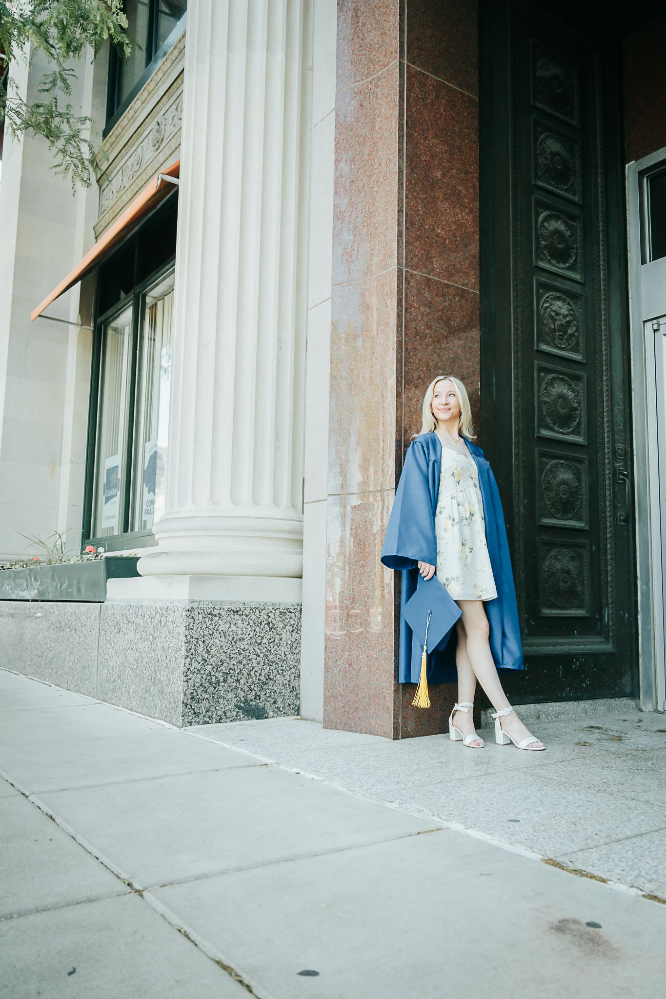 Blonde graduate in blue gown leaning against ornate building column holding cap with tassel.