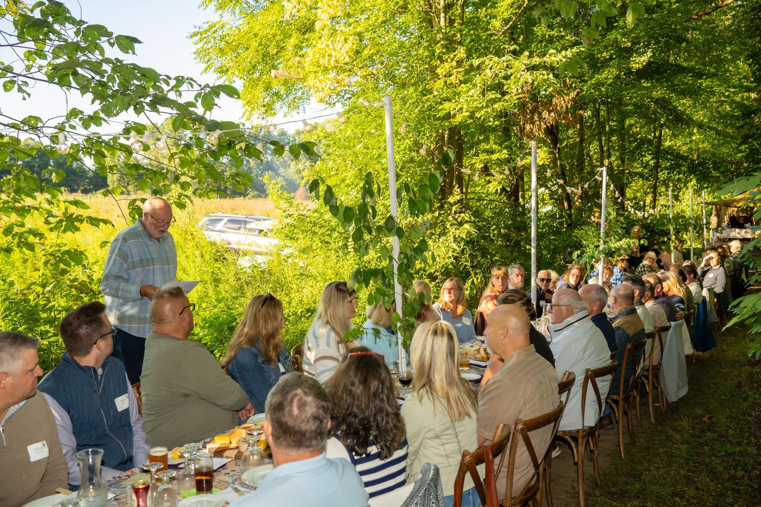 Large group of guests enjoying an outdoor woodland dinner party seated at long tables surrounded by green trees.