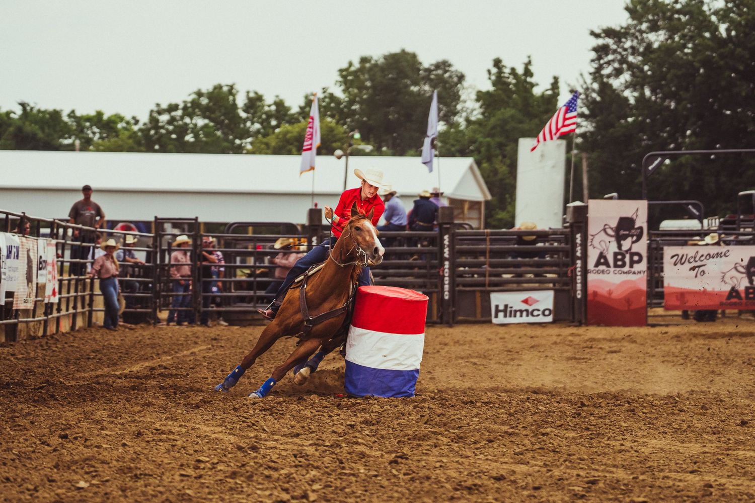 Barrel racing competitor on brown horse navigating red, white, and blue barrel at outdoor rodeo arena.