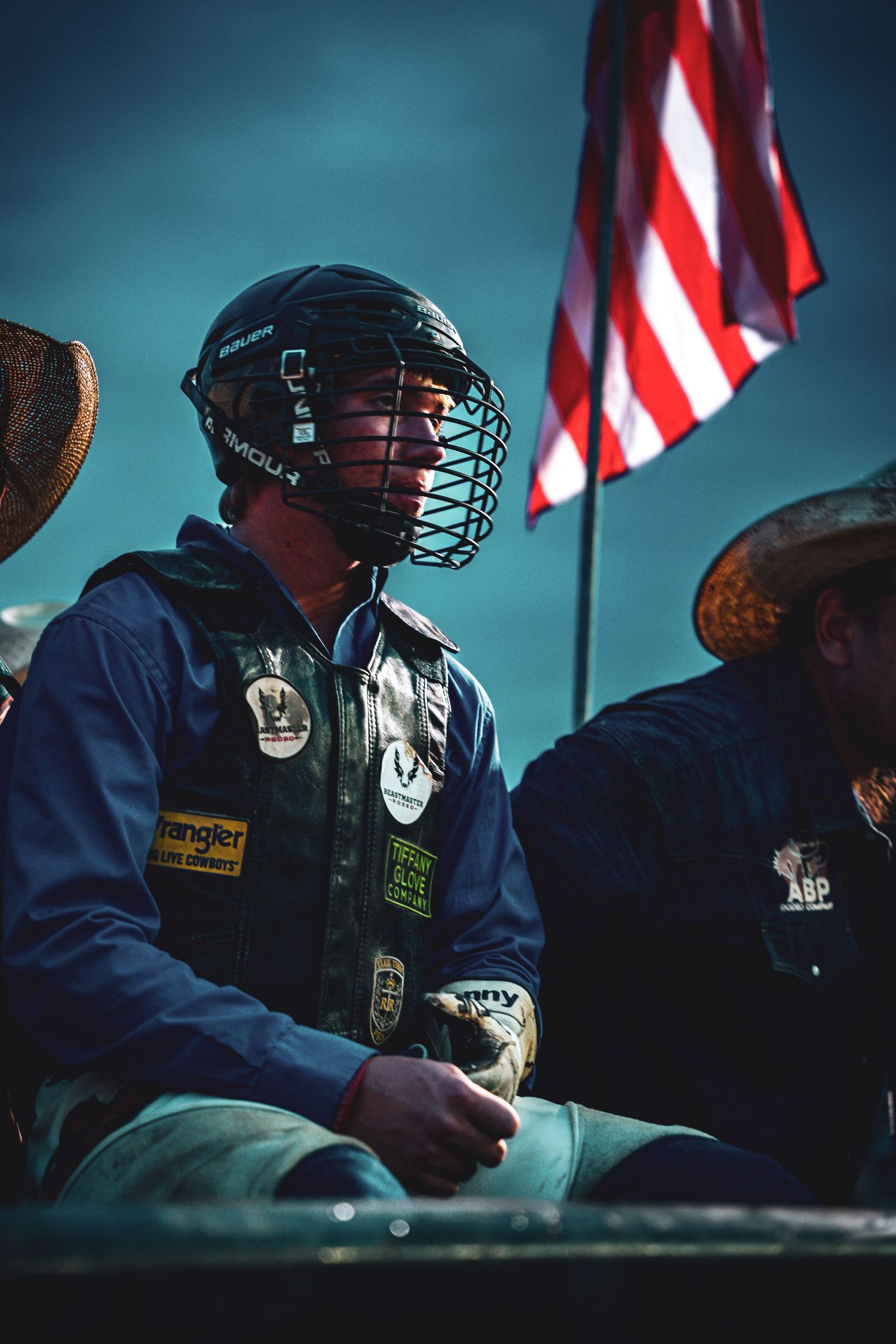 Bull rider in helmet and sponsor patches sits near an American flag at a rodeo event.
