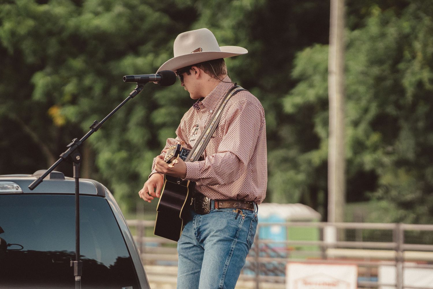 Young cowboy in hat playing acoustic guitar at outdoor microphone stand near rodeo arena.