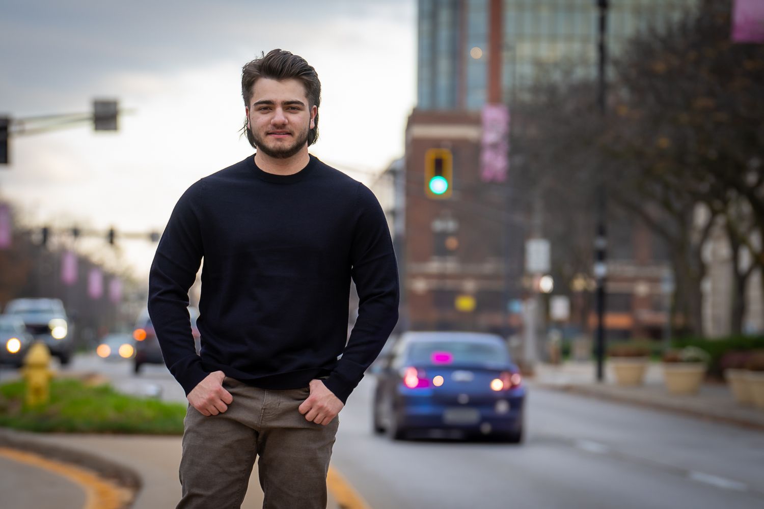 Young man in black sweater standing on urban street with traffic and city lights behind him.