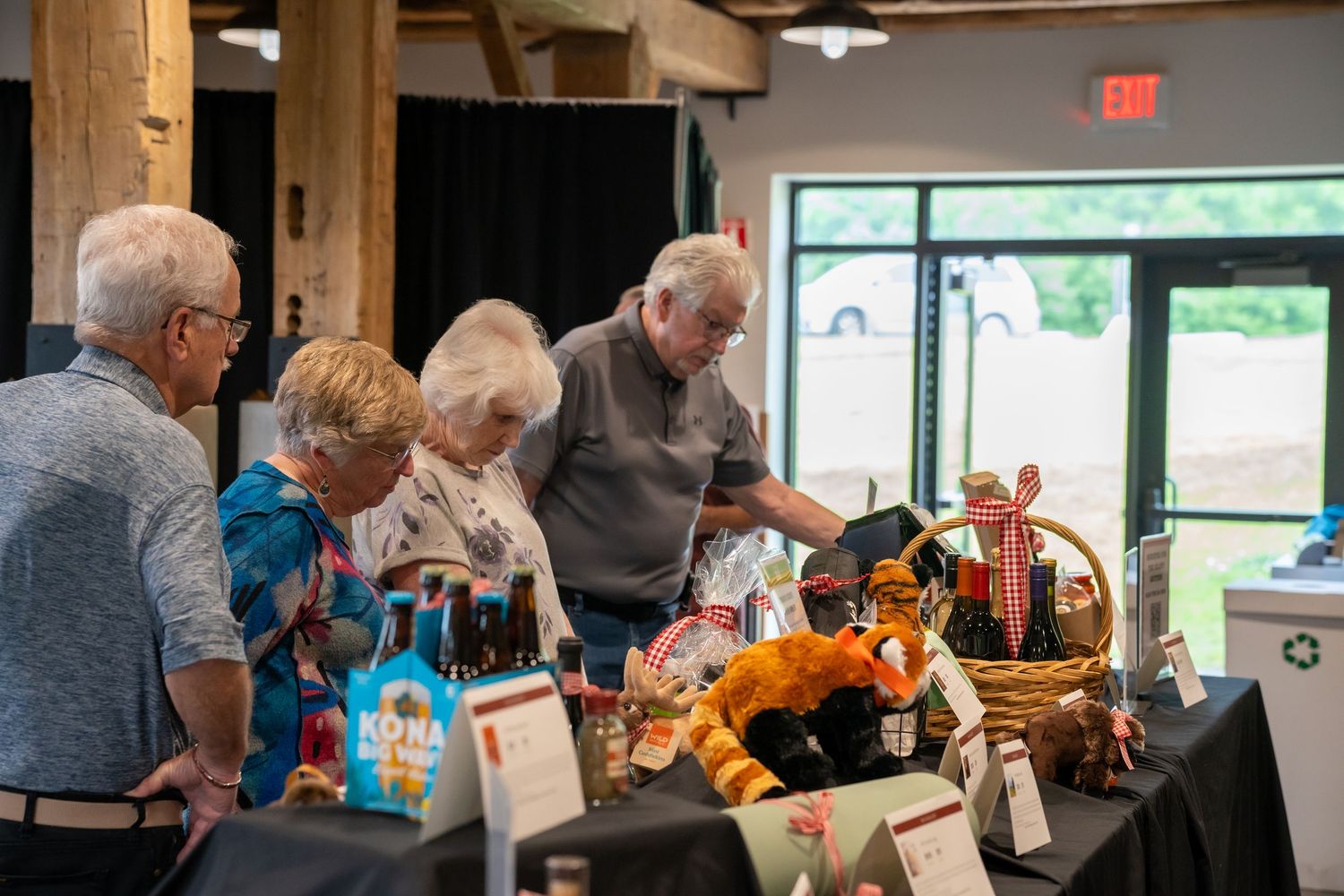 Elderly guests browsing silent auction gift baskets and prizes on a black draped table at an indoor fundraiser event.
