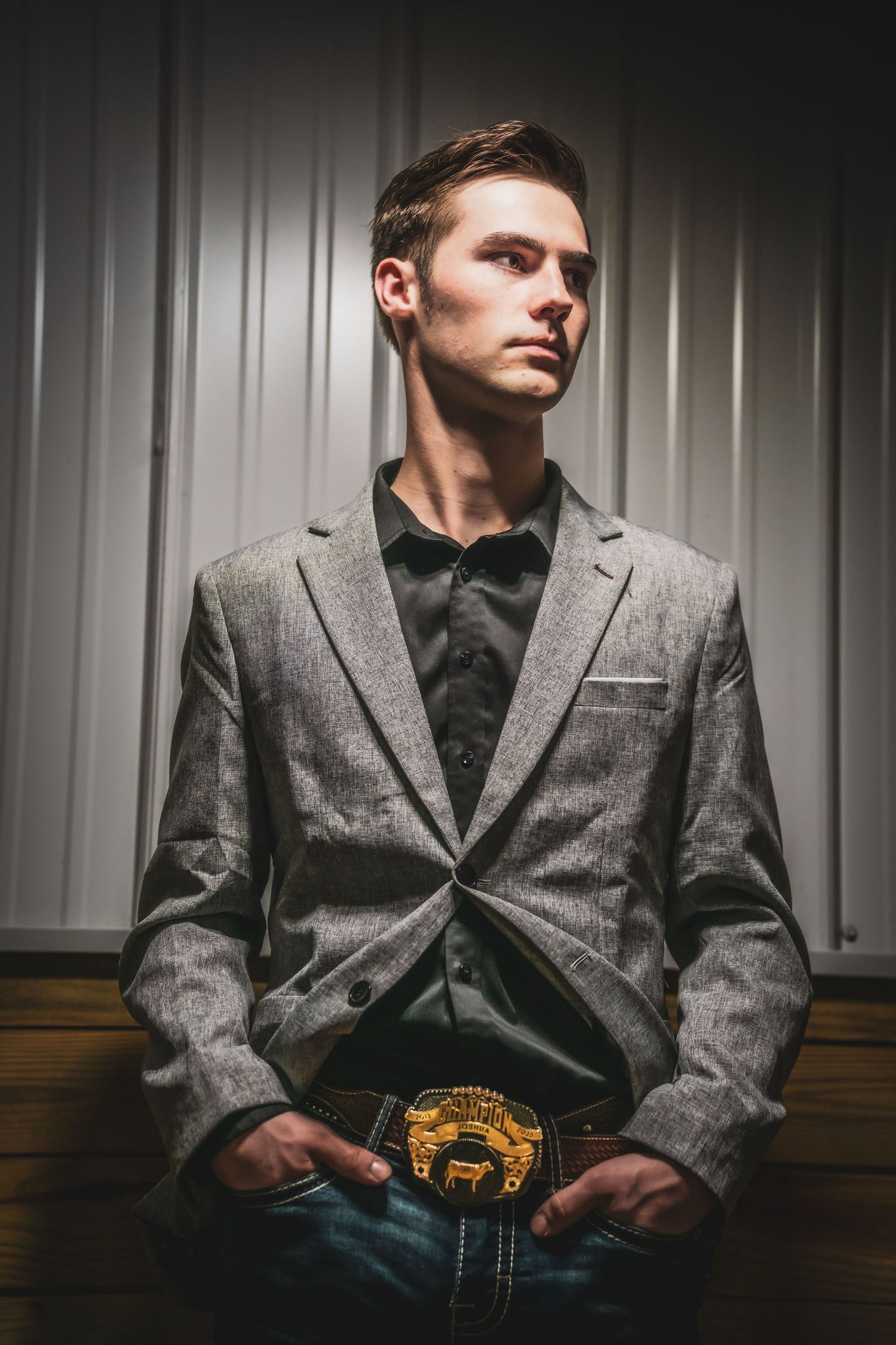 Young man in gray blazer and western belt buckle poses in dramatic low-key lighting.