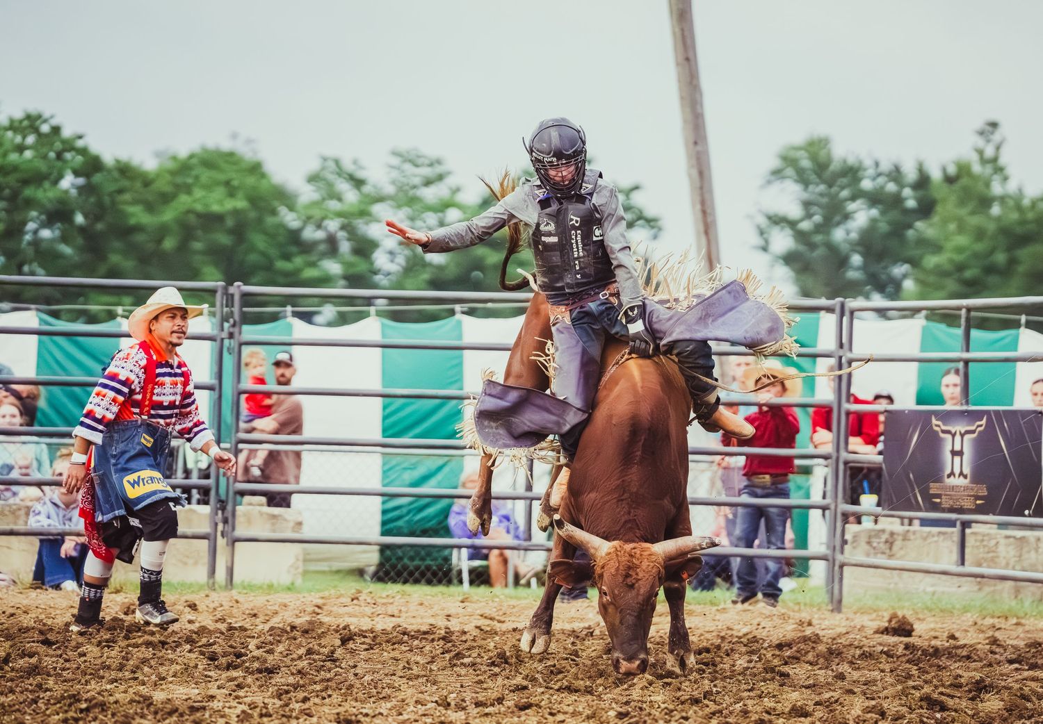 Rodeo rider on bucking horse with clown and crowd at outdoor dirt arena event.