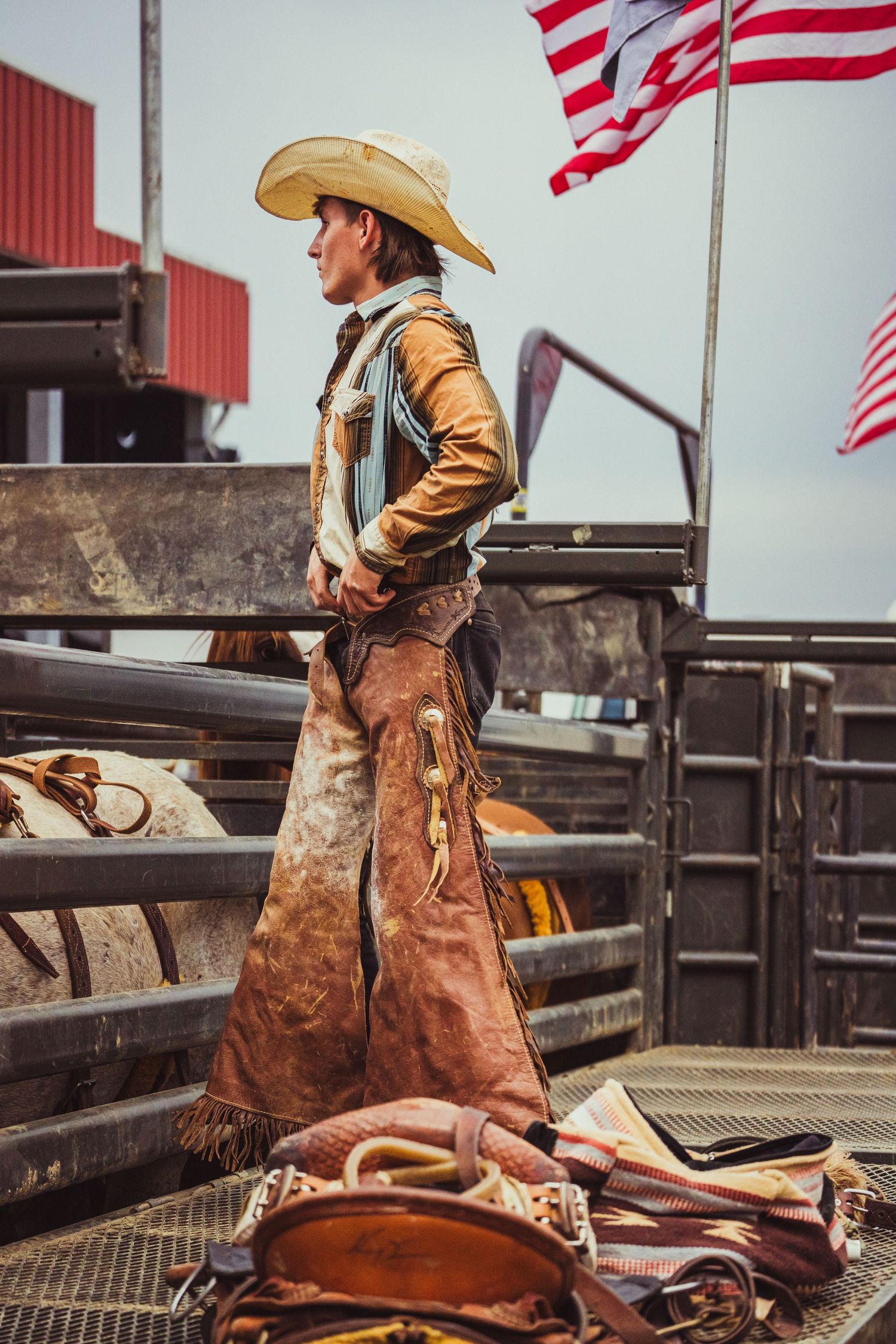 A cowboy in leather chaps and hat stands at a rodeo arena with an American flag waving in the background.
