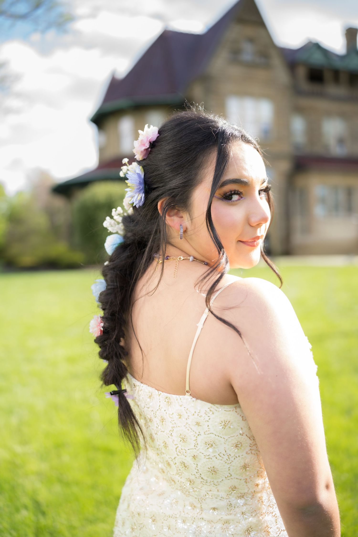 Young woman in floral braided hair and white dress poses outdoors near a historic stone mansion on a sunny day.