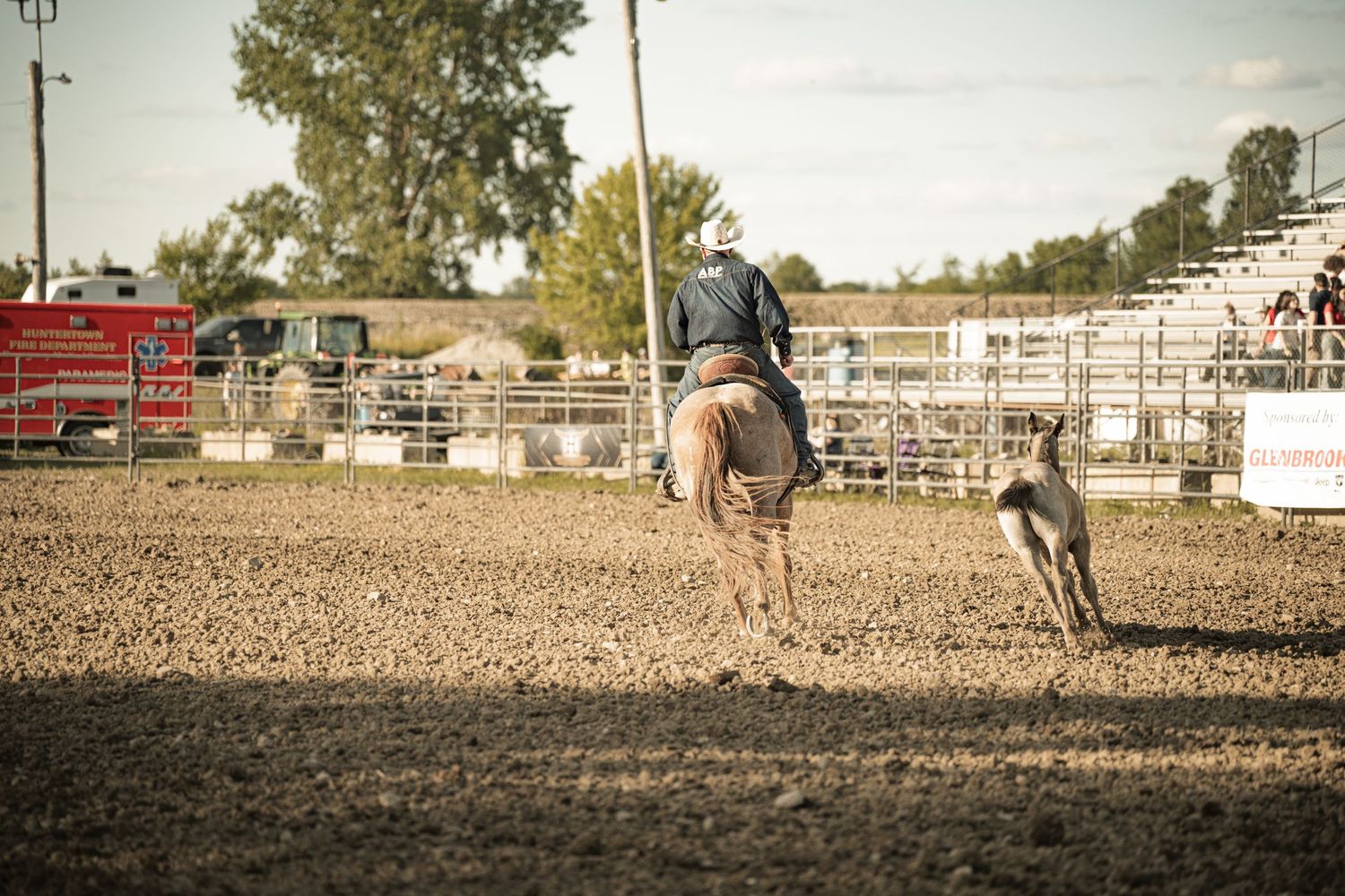 Cowboy on horseback chasing a calf across a dirt arena during a rodeo competition.