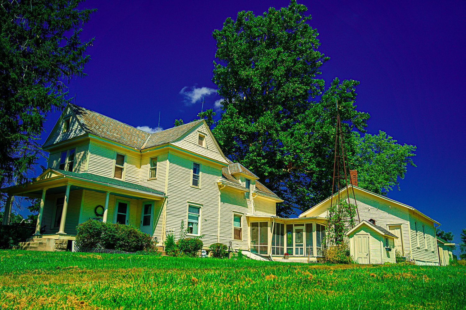 A charming yellow Victorian farmhouse with wraparound porch surrounded by green trees under deep blue sky.
