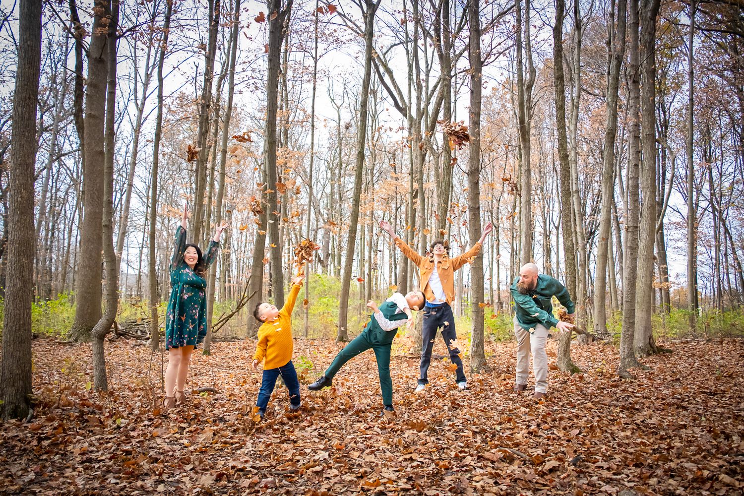 A joyful family tosses autumn leaves in a colorful fall forest with tall bare trees.