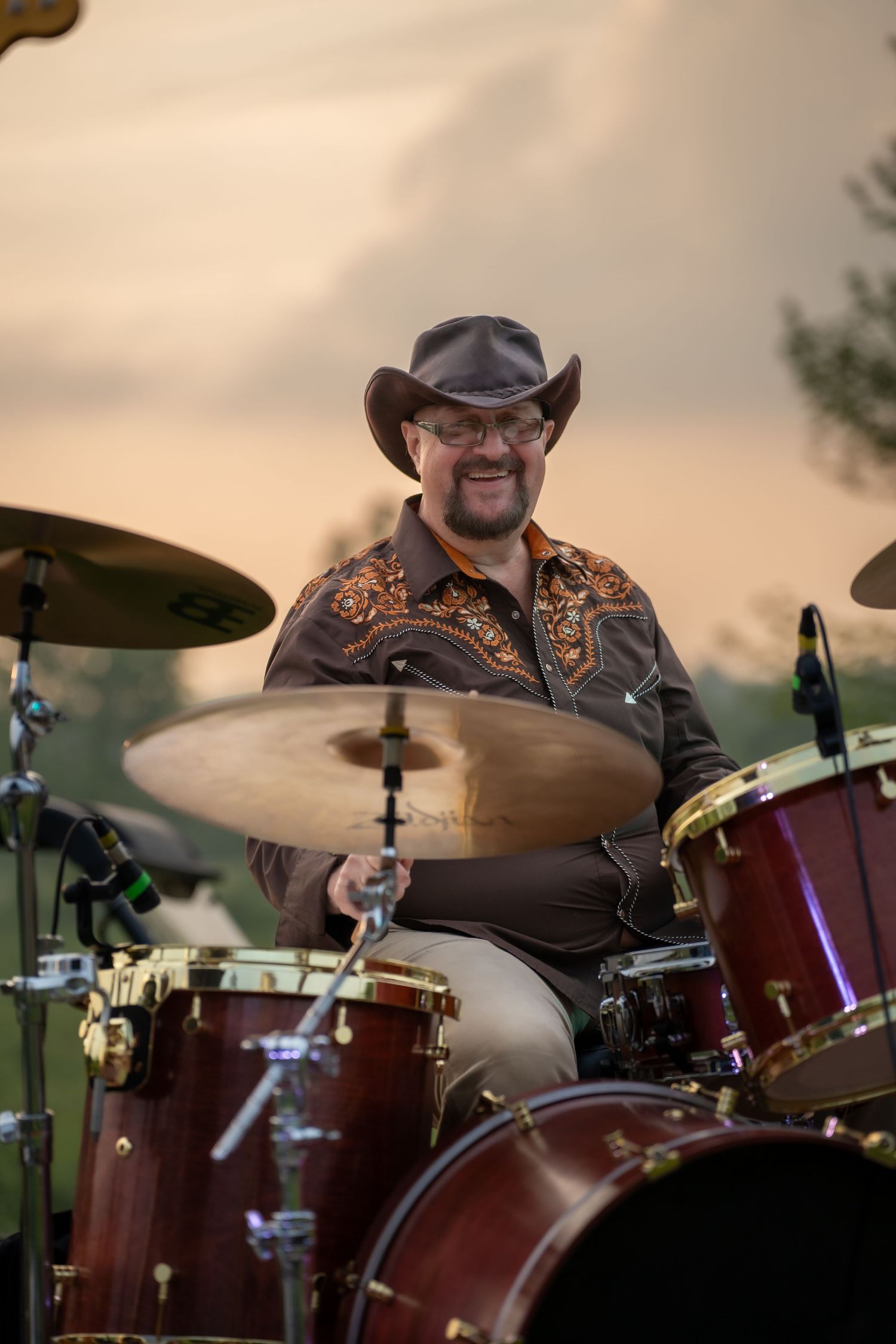 Smiling drummer in cowboy hat playing red drum kit at outdoor sunset concert.