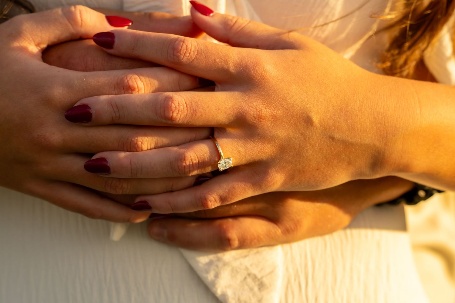 Close-up of an engagement ring on a woman's hand with red nails resting on her partner's hands.