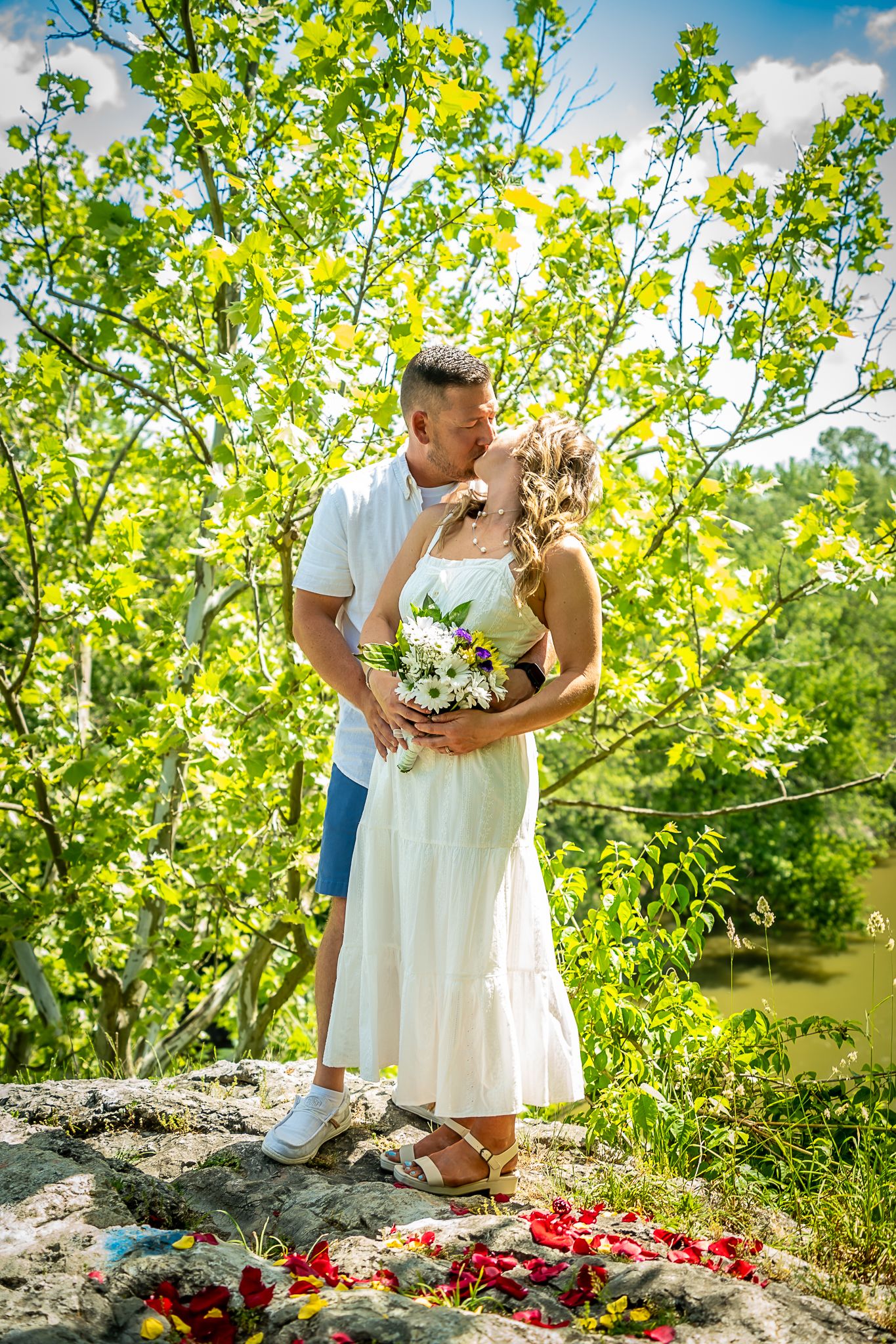 Couple kissing on rocky overlook surrounded by lush green trees, woman holding wildflower bouquet.
