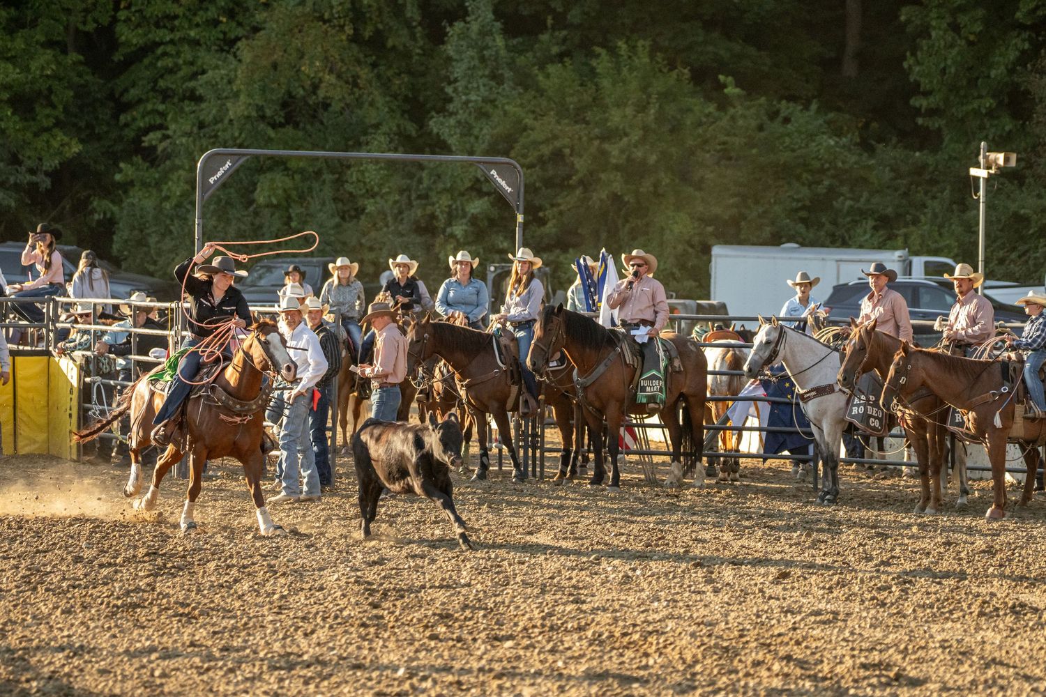 Cowboys on horseback practice roping a calf in a dusty rodeo arena surrounded by other riders.