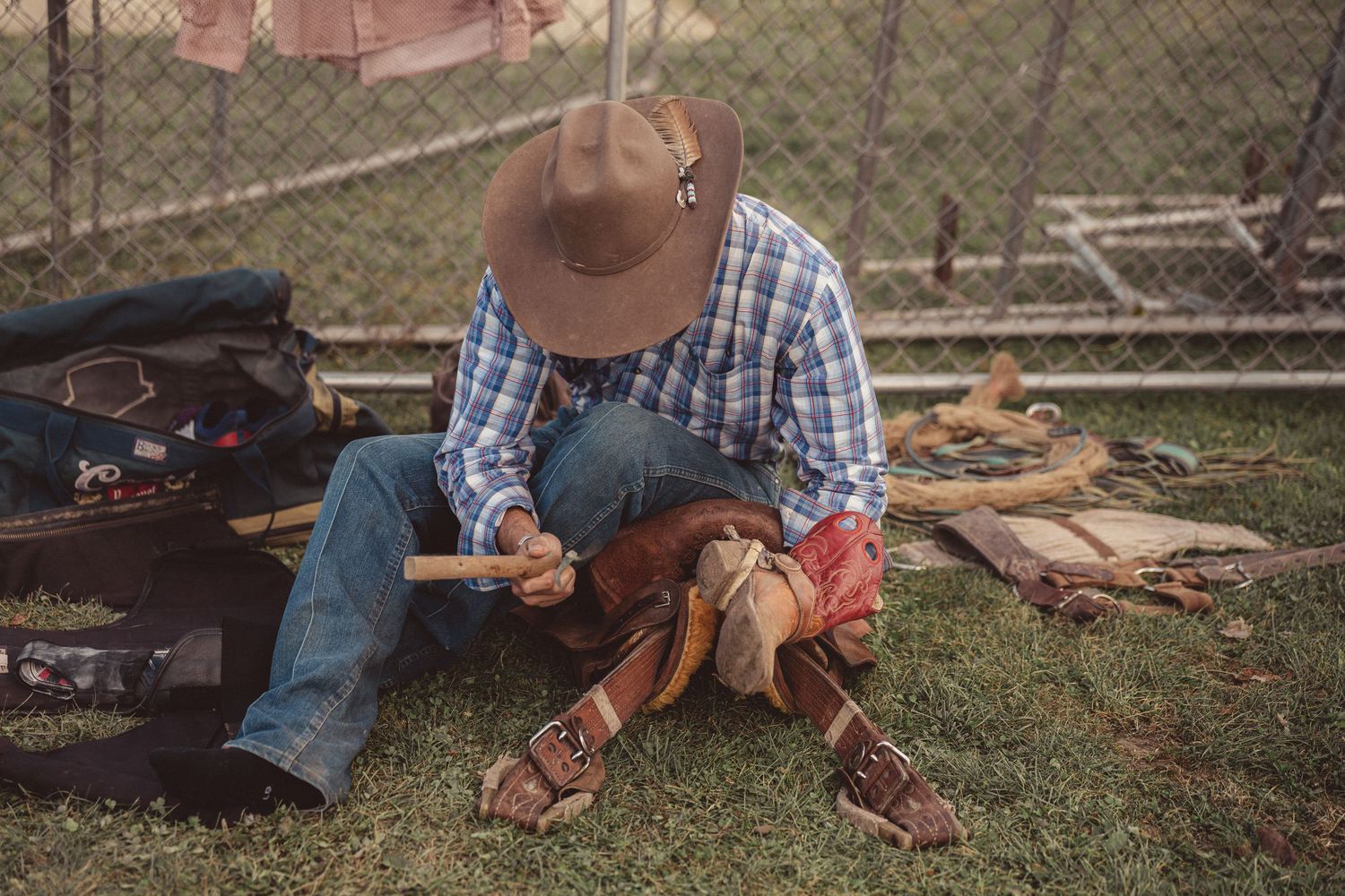 Cowboy in plaid shirt and brown hat working on leather saddle equipment while sitting on ground.