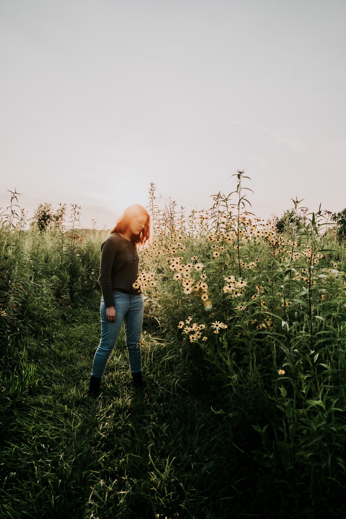 A red-haired woman in jeans and dark top stands among tall wildflowers in a green meadow at golden hour.