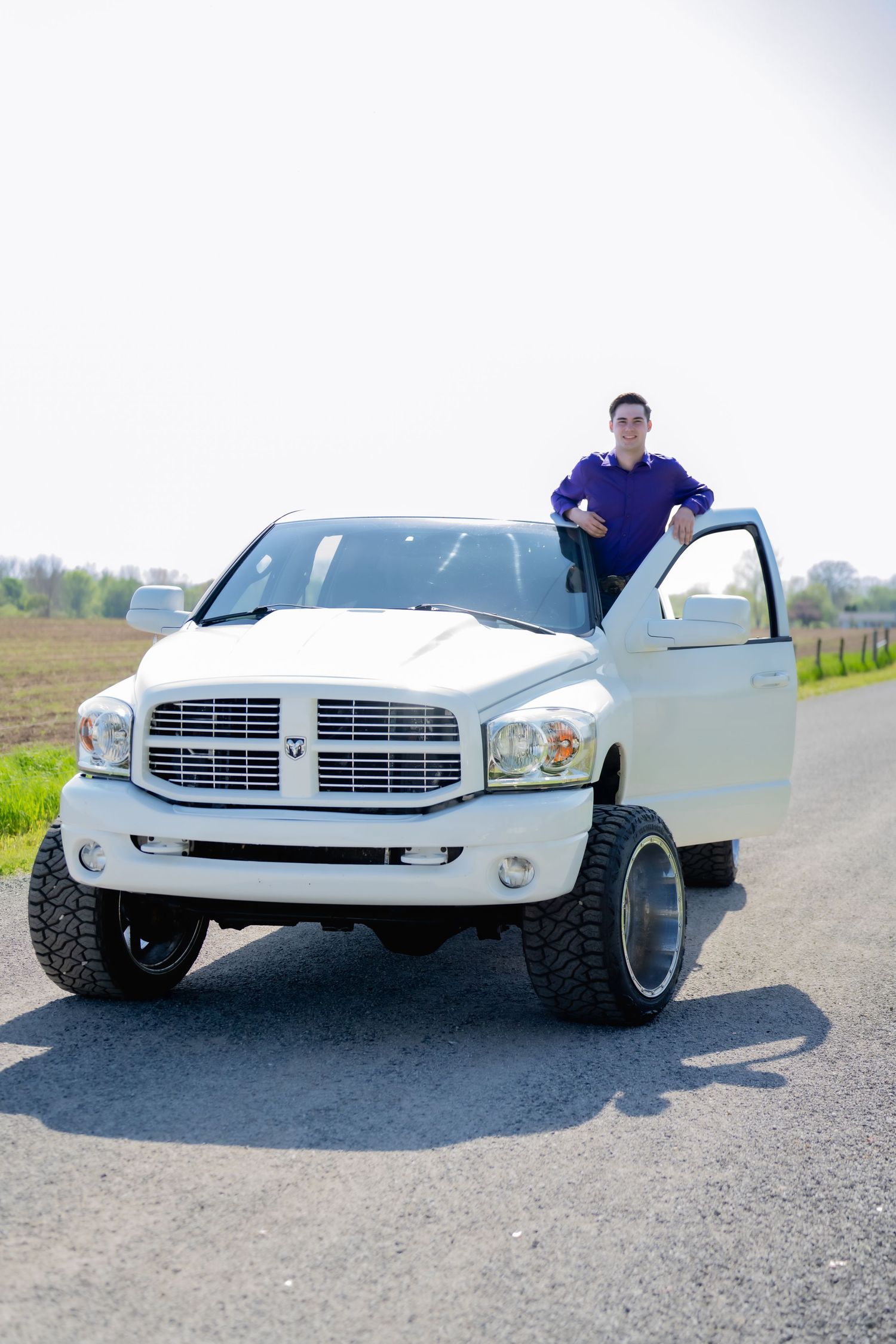 Young man in purple shirt standing beside lifted white Dodge Ram truck on a rural country road.