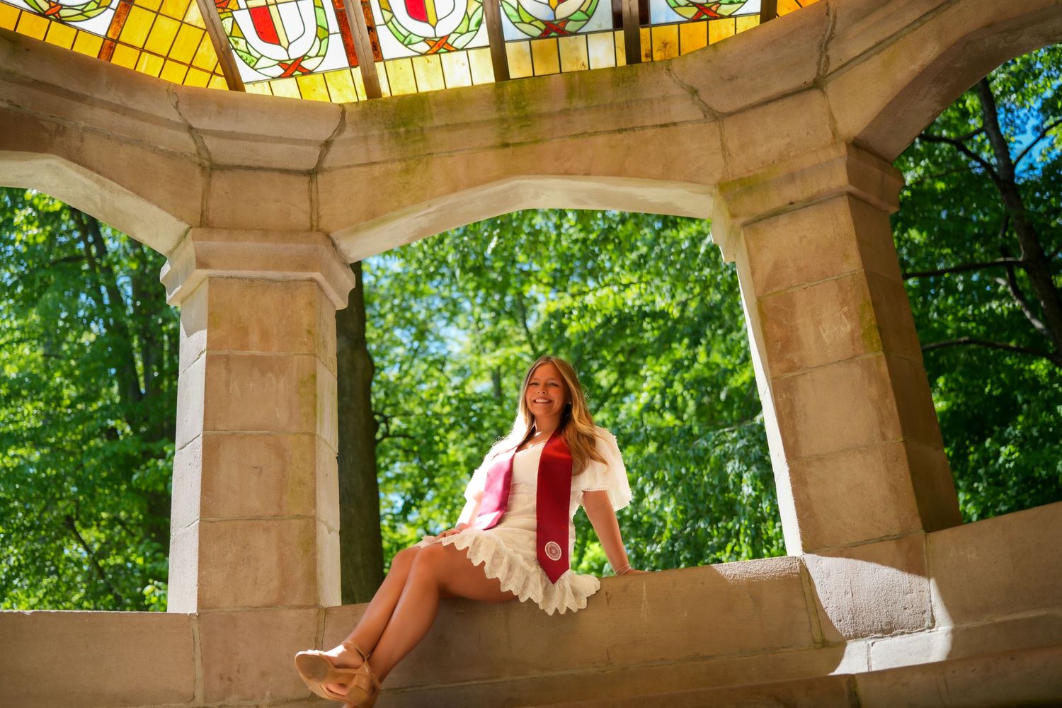 Smiling graduate in white dress and red stole sitting in ornate stone gazebo with stained glass ceiling.
