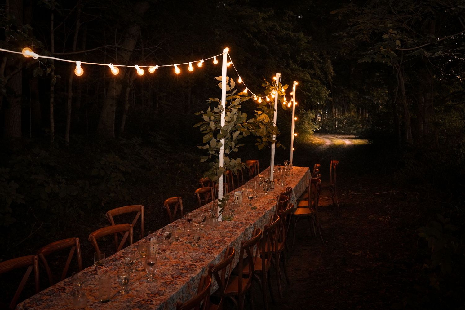 Long rustic outdoor dining table lit by string lights and tall candles in a dark forest setting at night.