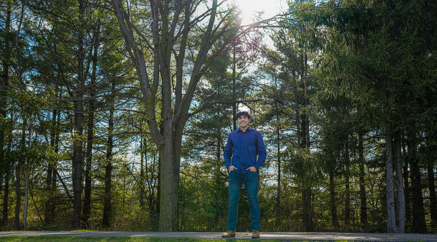 Man in blue jacket and white hat standing on path surrounded by tall green trees in sunlit park.