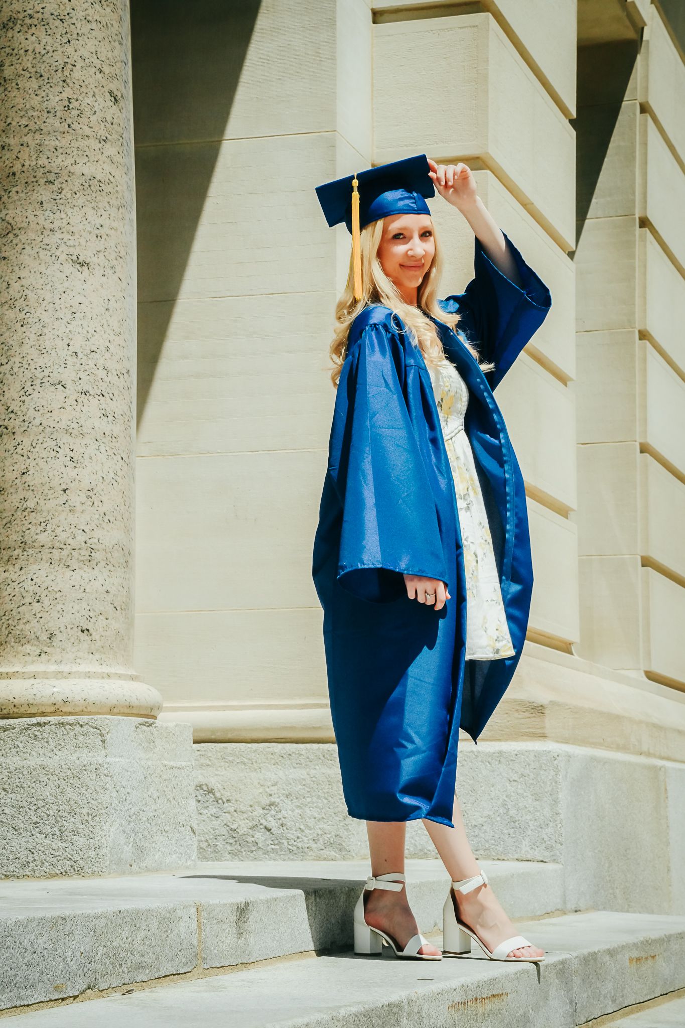 Blonde graduate in blue cap and gown posing on steps of classical building with columns.