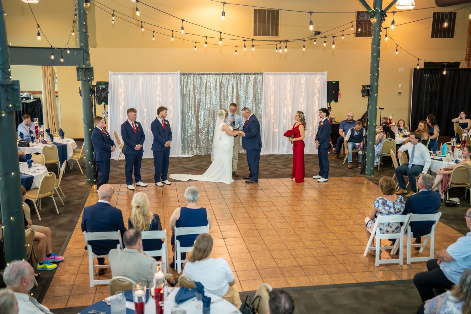 Wedding ceremony indoors with string lights, groomsmen in navy suits, bride in white, and maid of honor in red dress.