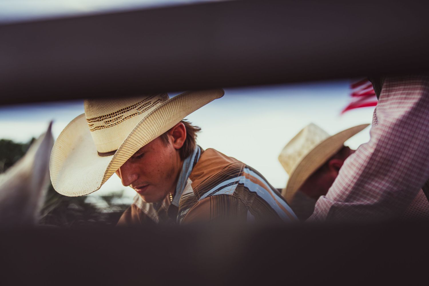 Two cowboys wearing straw hats at a rodeo event, photographed through metal rails in warm natural light.