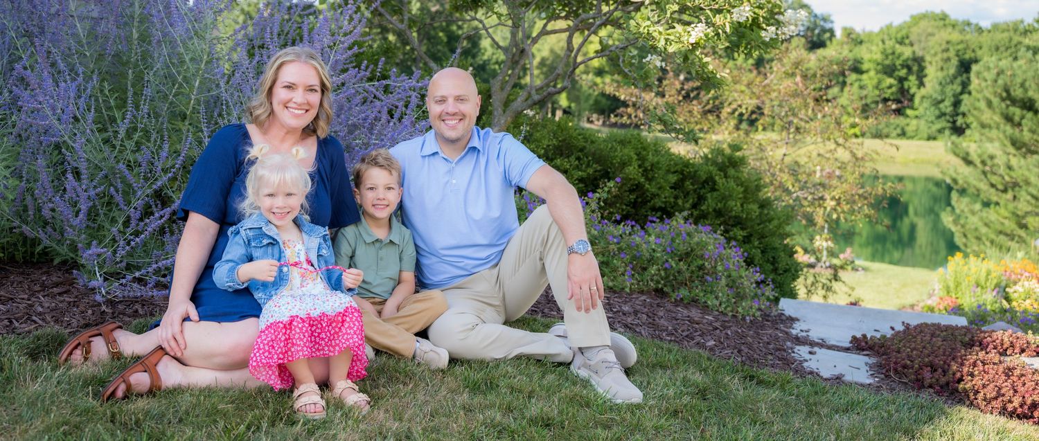 Family of four smiling together on lush green grass surrounded by purple flowers and scenic pond backdrop.