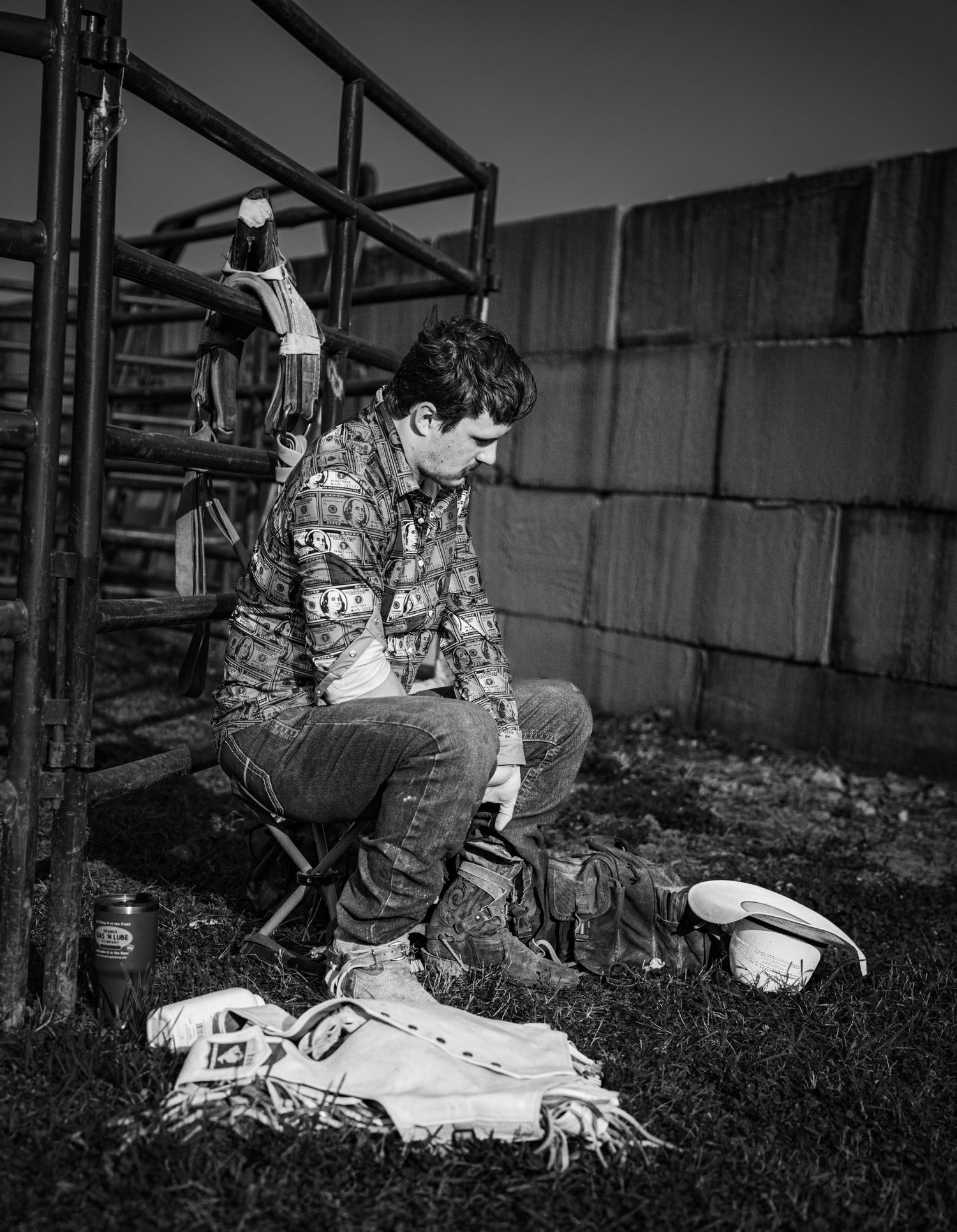 Black and white photo of a young man sitting on a stool near metal gates, looking down at gear on the grass.