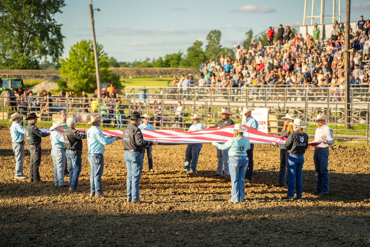 Cowboys hold a large American flag during a rodeo opening ceremony before a packed grandstand audience.