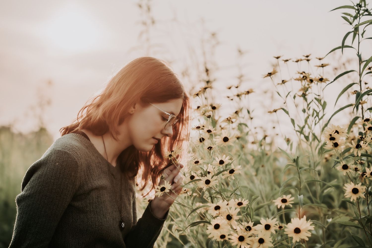 Young woman with glasses smelling wildflowers in a field at golden hour, surrounded by black-eyed Susans.