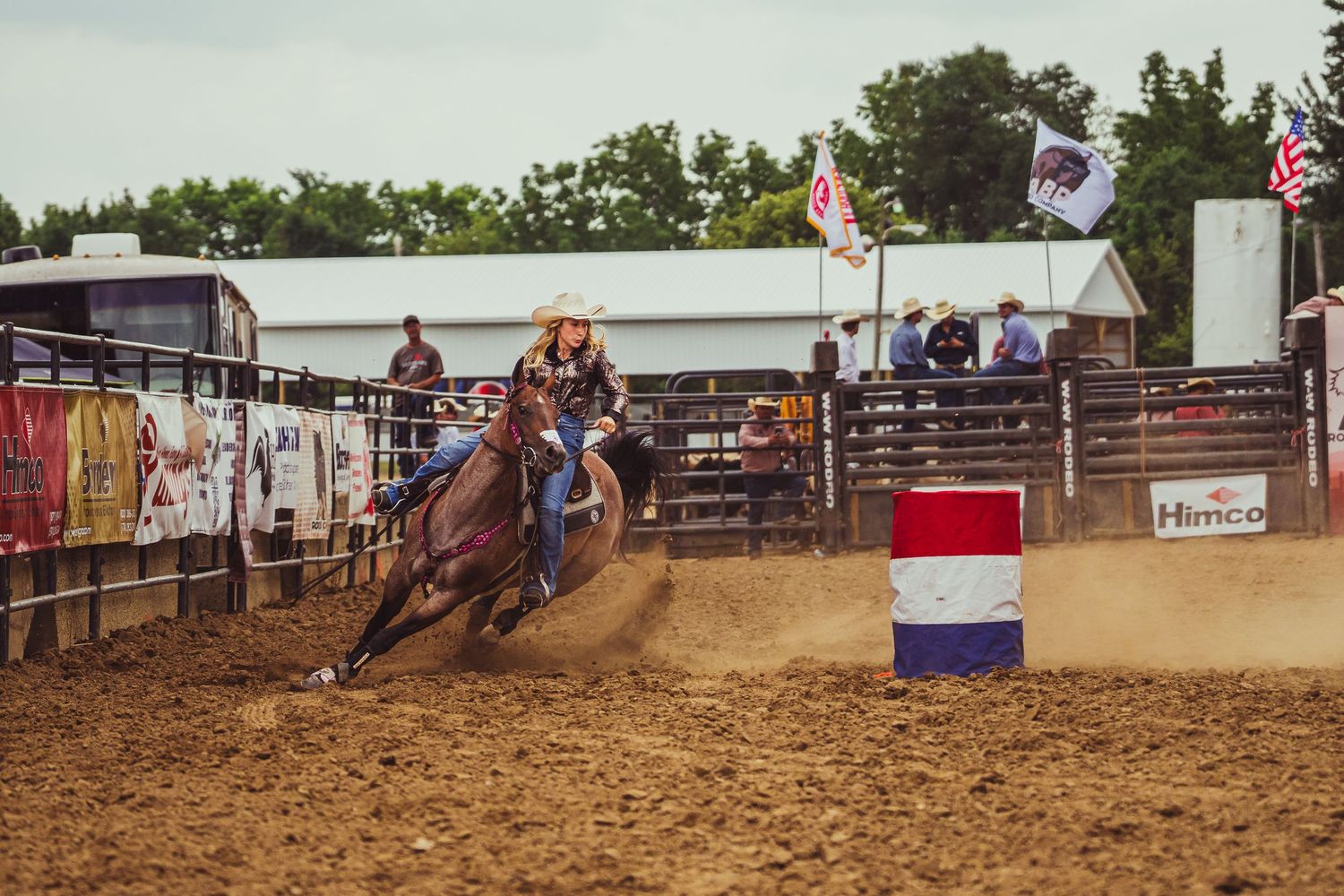 Cowgirl riding horse around barrel in dusty rodeo arena during barrel racing competition.