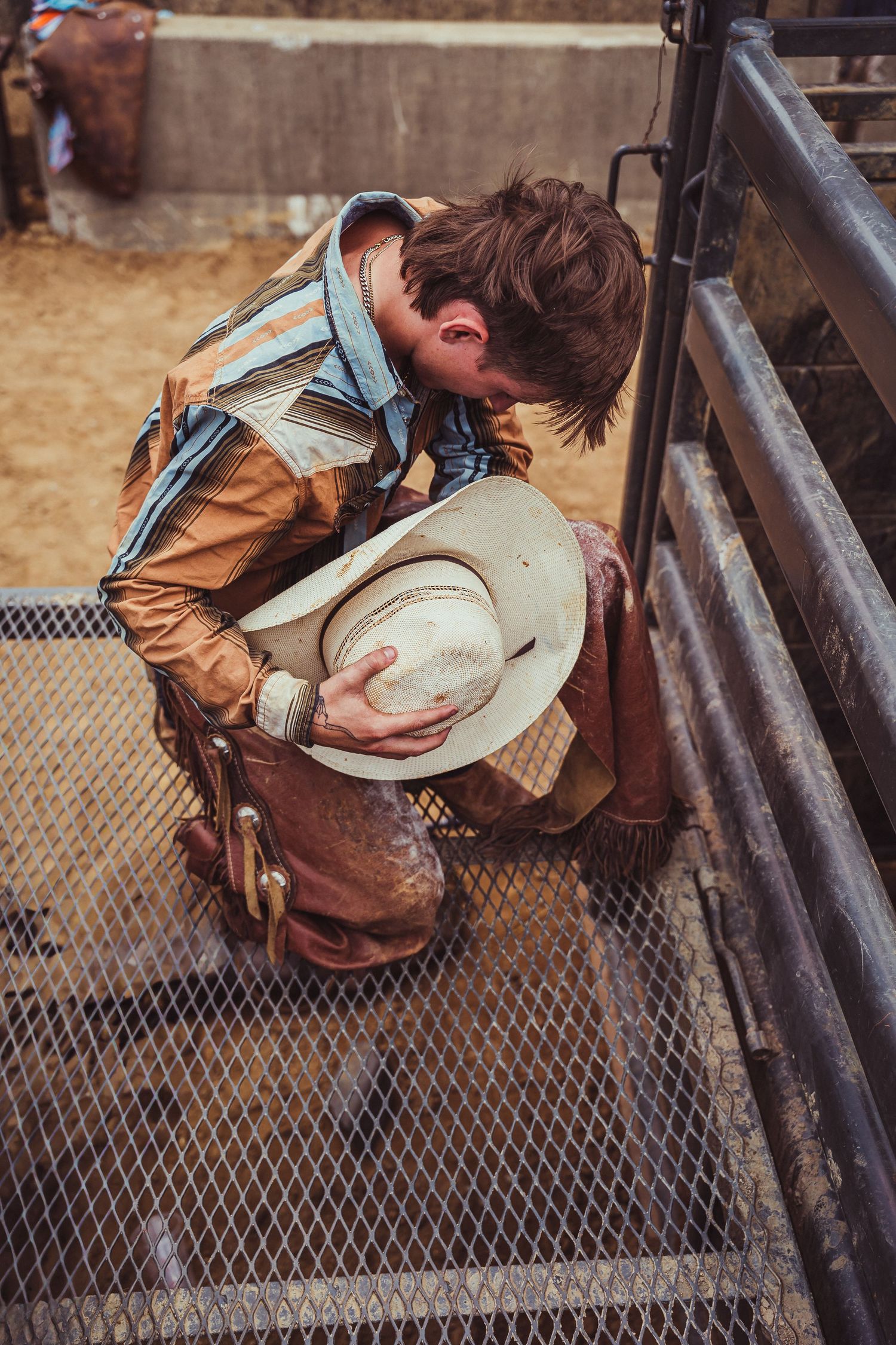 A cowboy kneels in prayer holding his white hat at a rodeo arena, wearing leather chaps and a blue shirt.