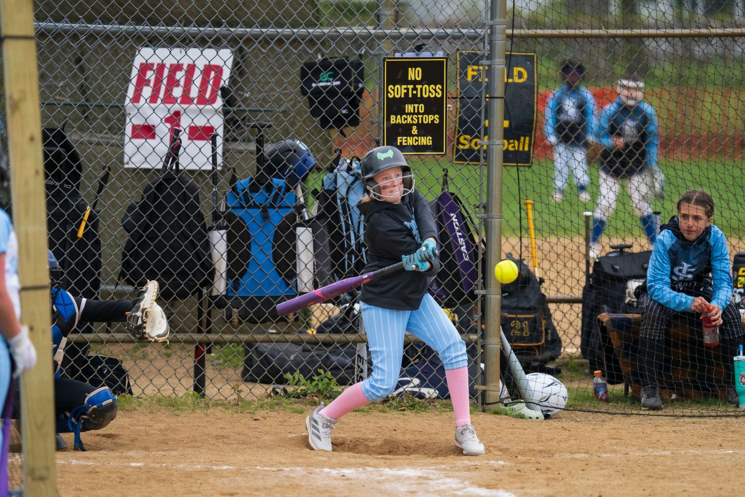 Young girl batting in a youth softball game at a fenced field, wearing a helmet and swinging at a pitch.