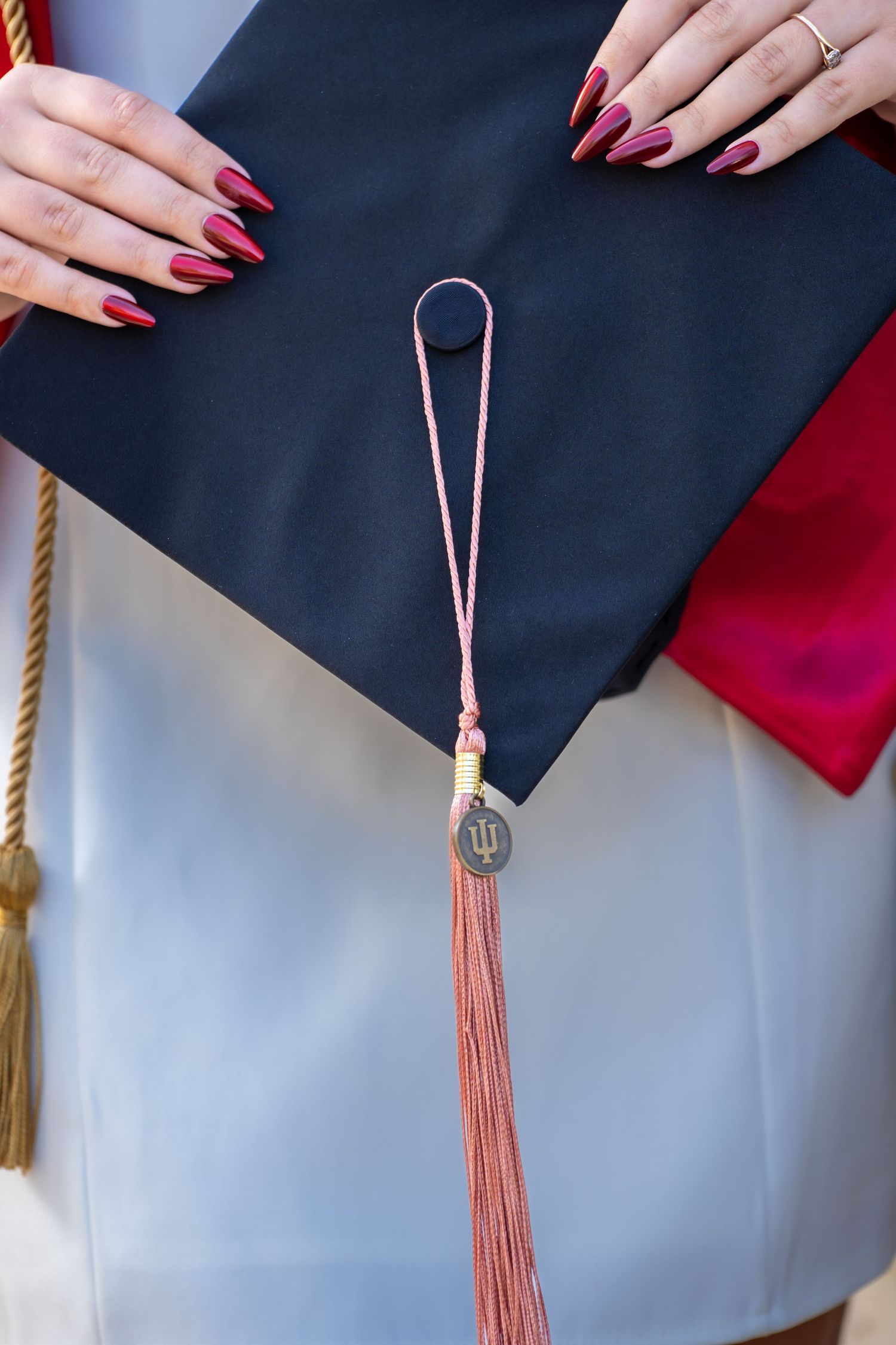 Close-up of graduation cap with pink tassel and IU medallion, held by hands with dark red manicured nails.