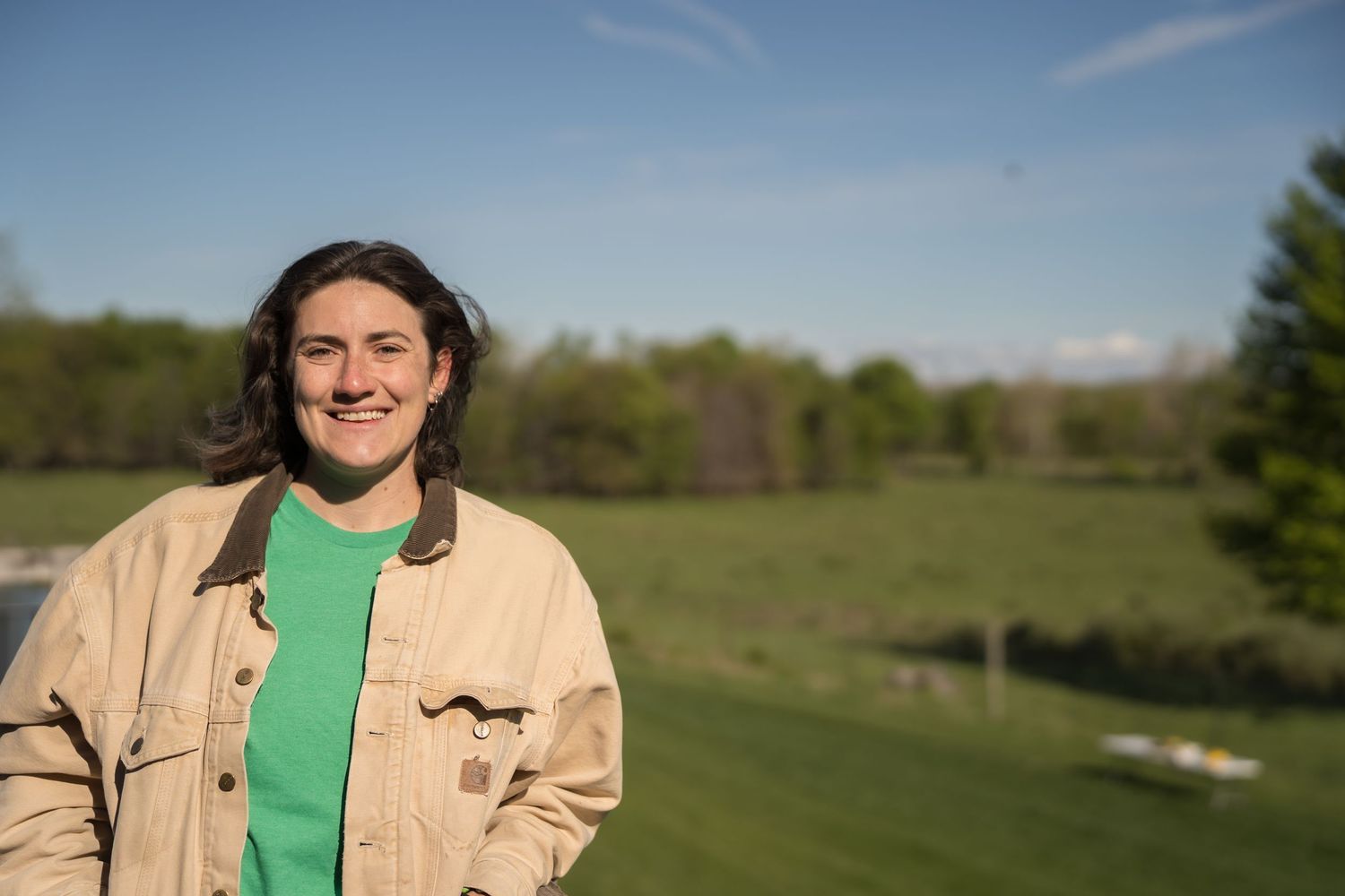 Smiling young woman in beige jacket and green shirt standing outdoors in a lush green field on a sunny day.