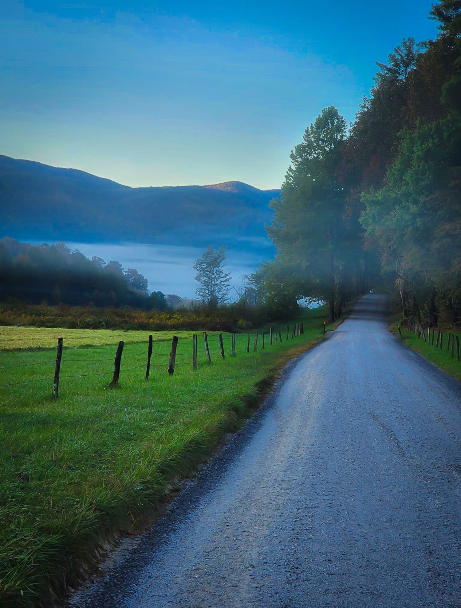 Scenic gravel road with wooden fence leading through misty mountain valley at blue hour dawn.