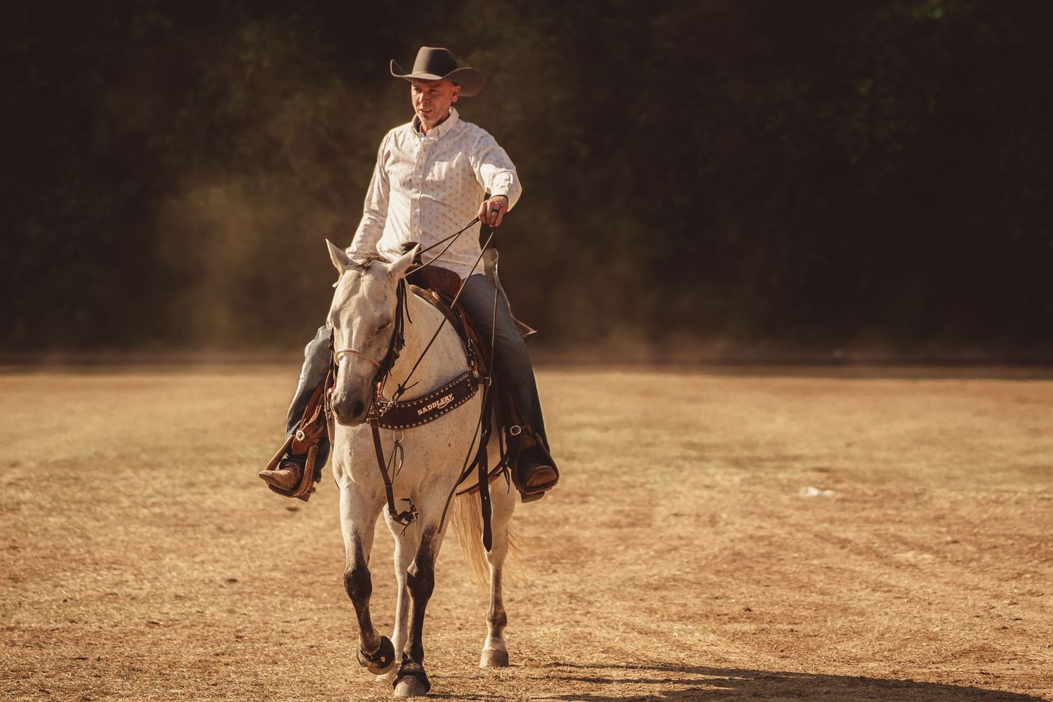 Cowboy riding white horse across dusty field in golden sunset light at outdoor ranch.