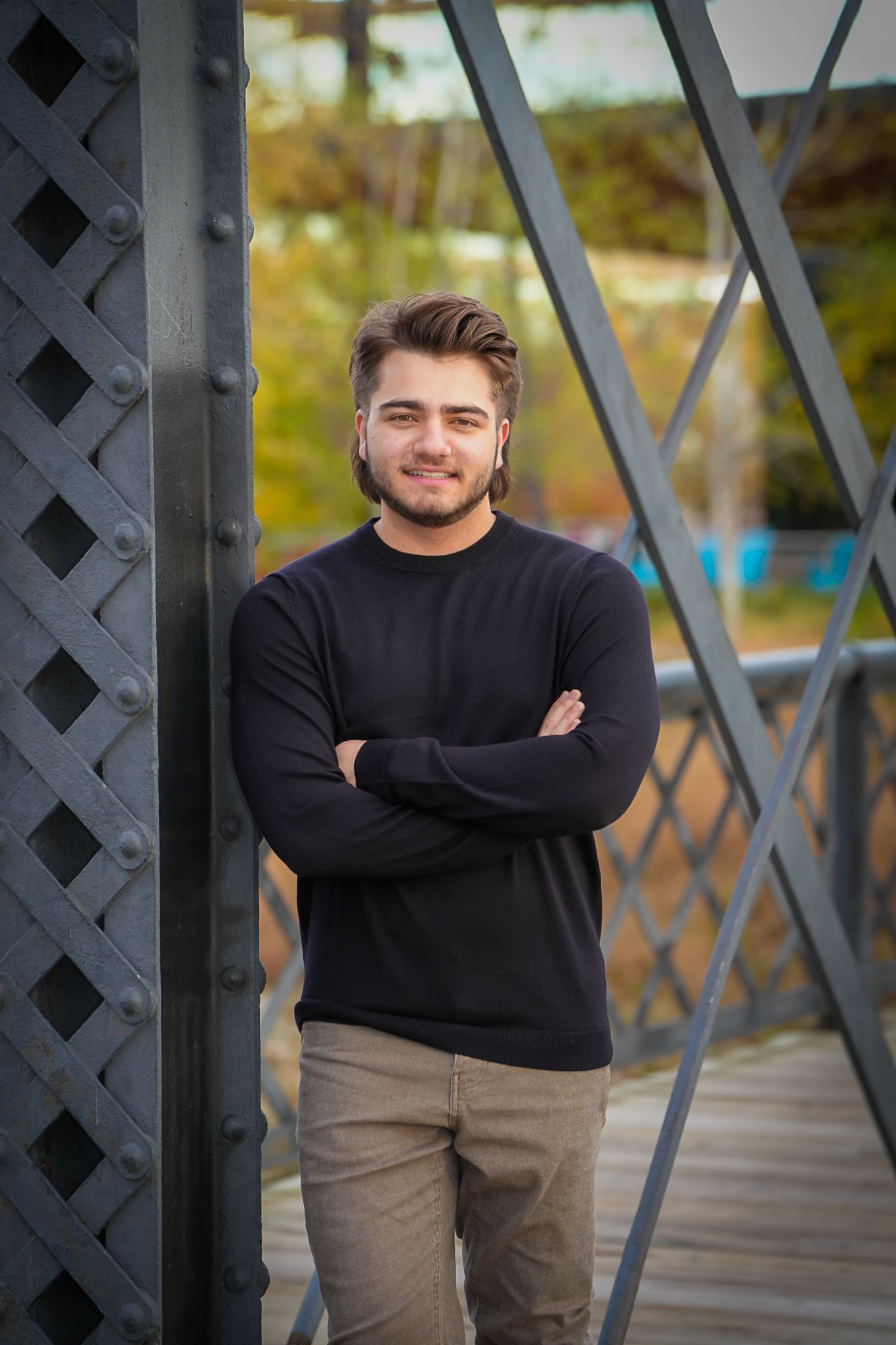 Young bearded man in black sweater with arms crossed leaning against metal bridge structure.