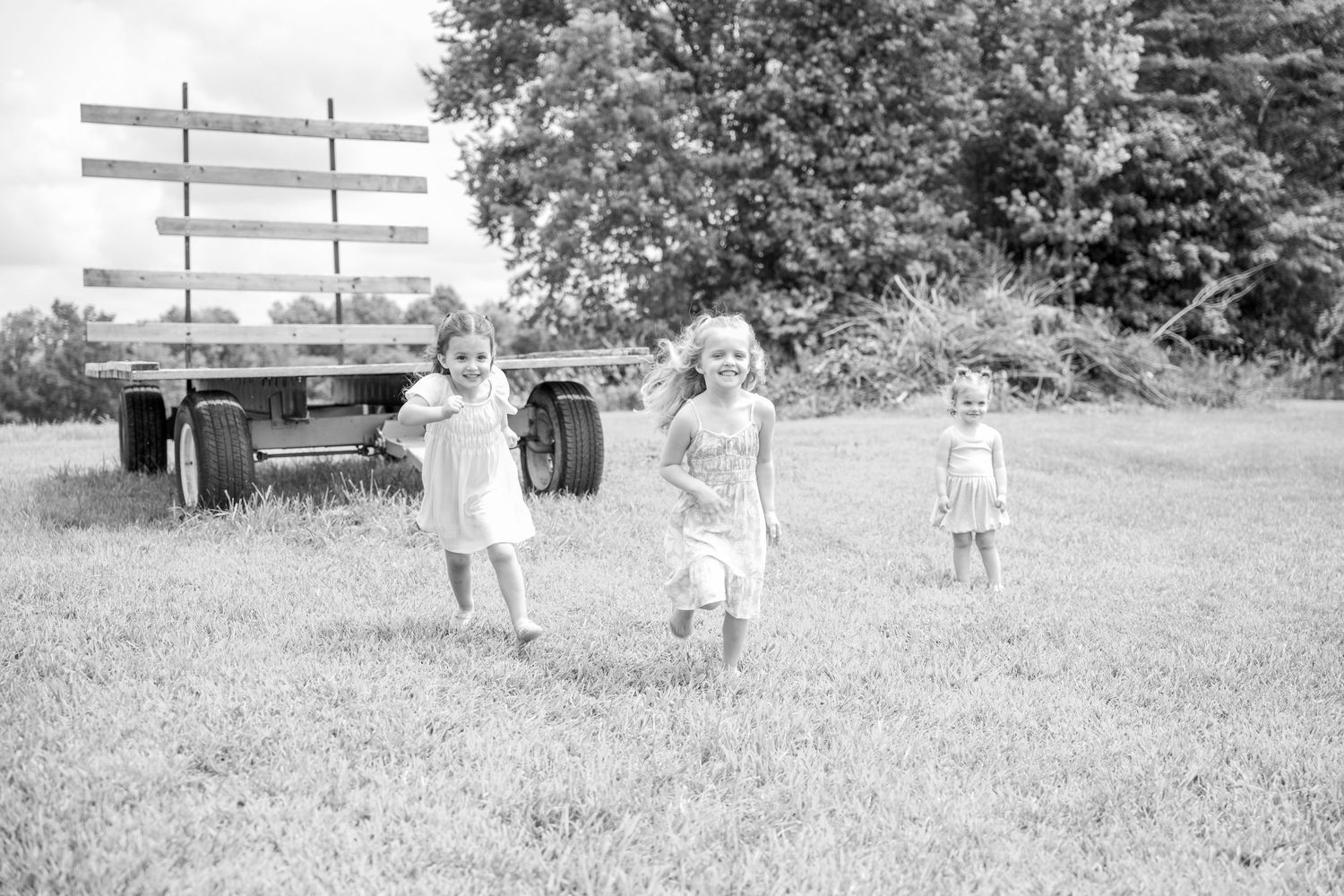 Three young girls running and playing in a grassy field beside a wooden farm wagon in a vintage black and white photo.