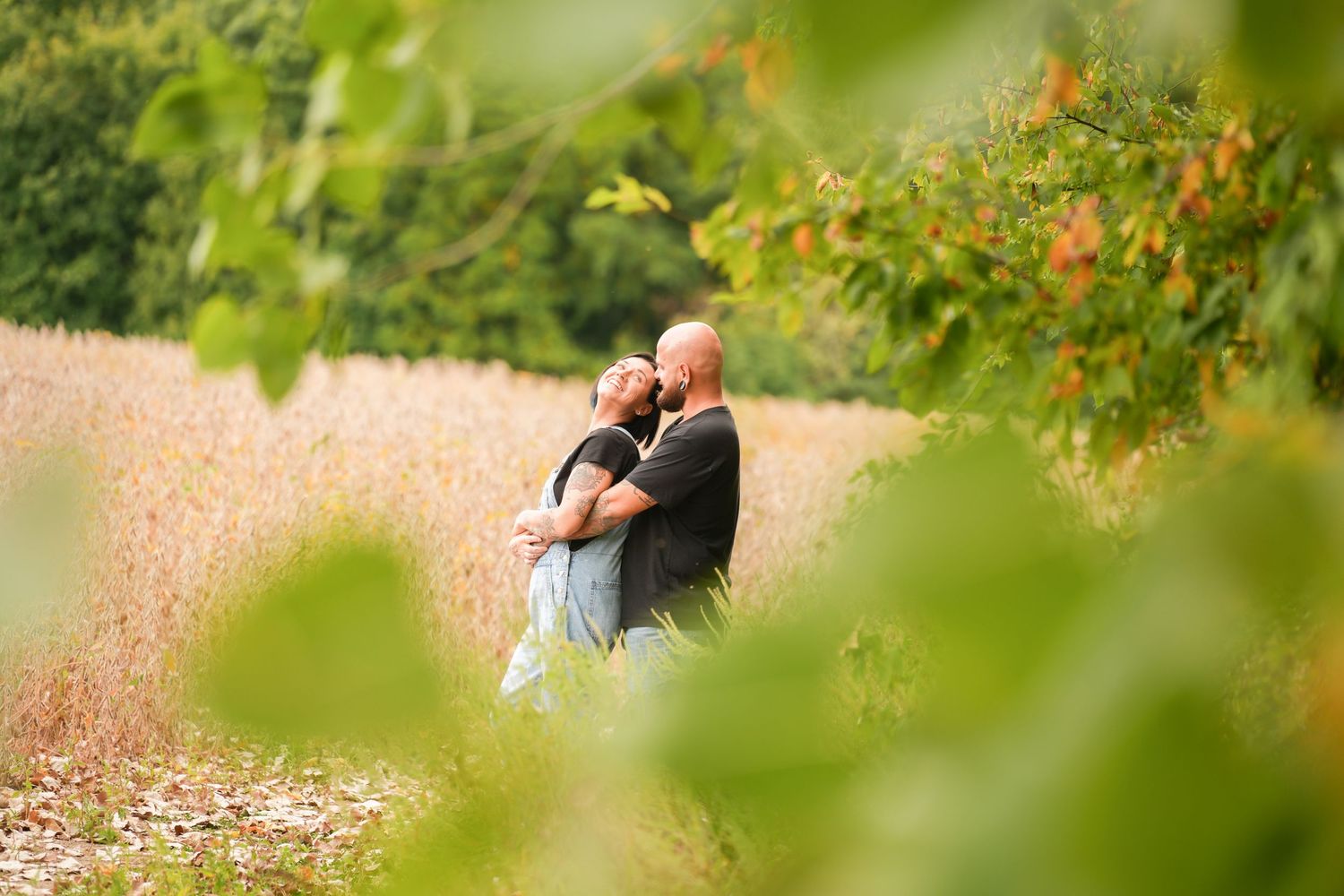 Couple embracing romantically in a lush green field surrounded by autumn foliage and tall golden grasses.