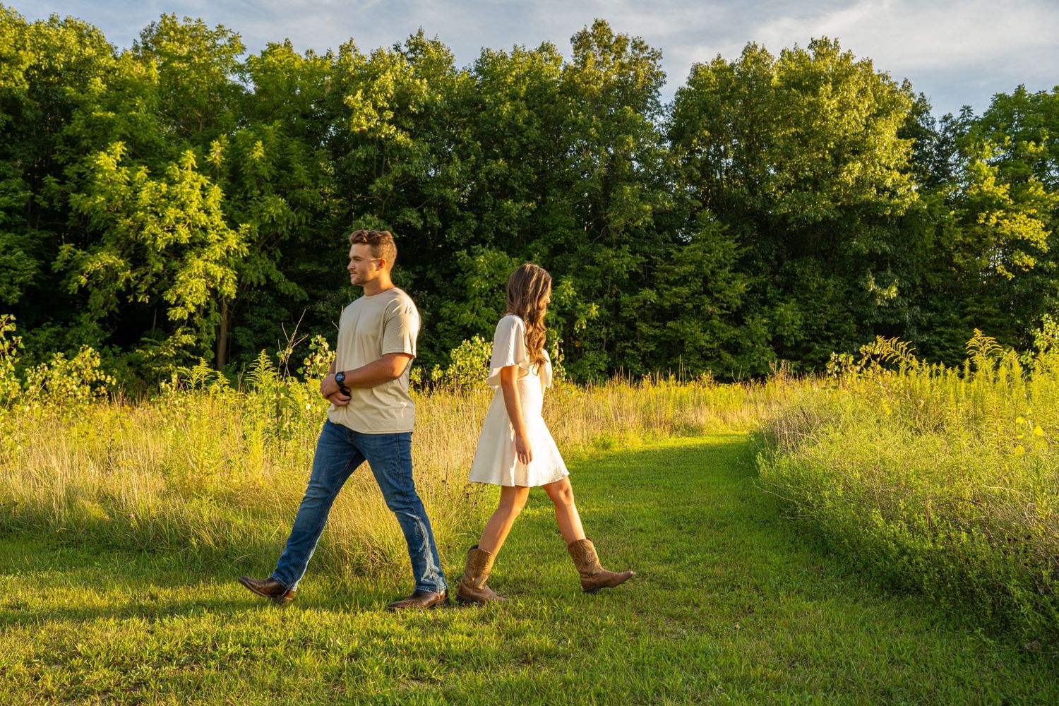 Couple walking hand-in-hand along a sunny green meadow path surrounded by lush trees.