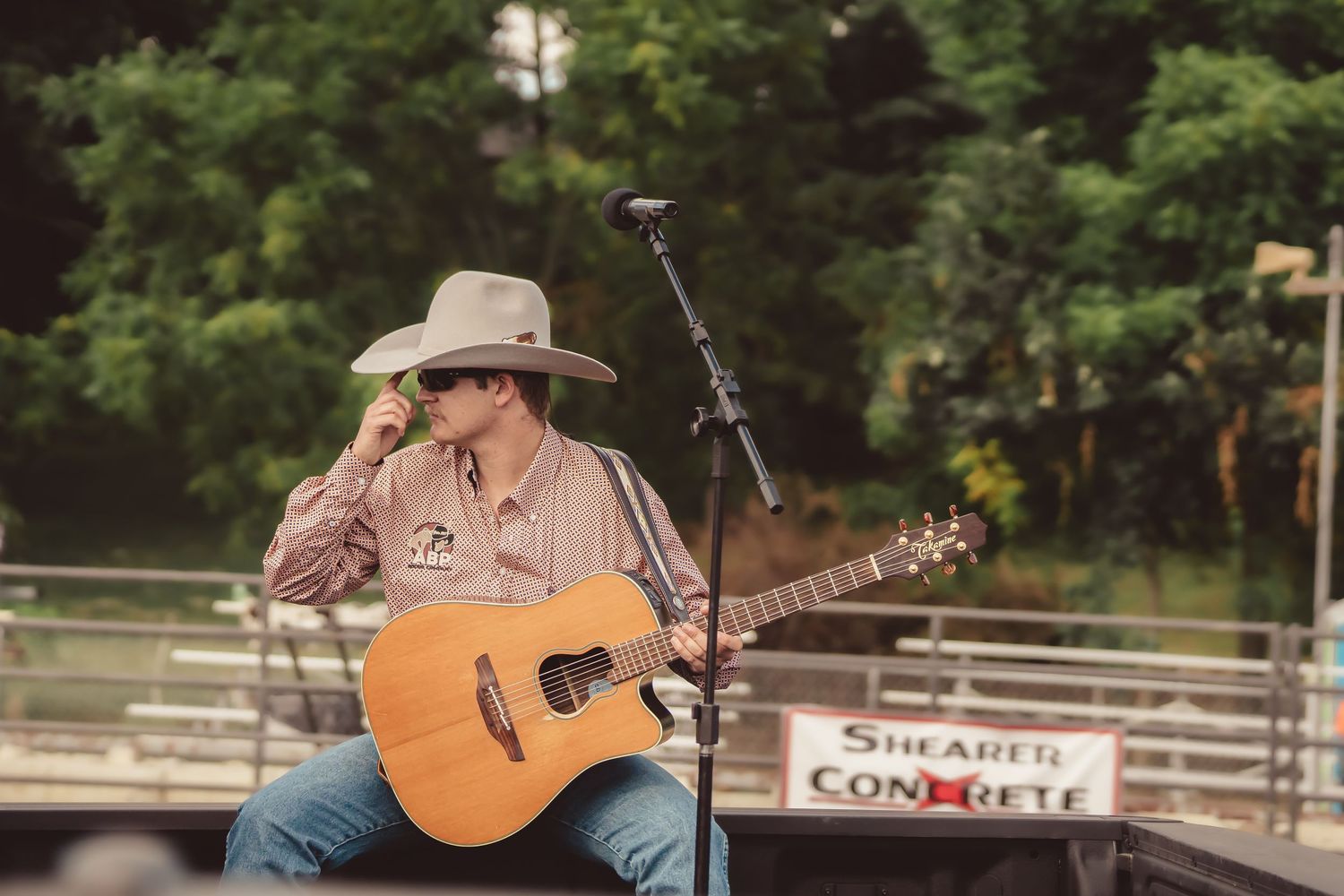 Cowboy musician playing acoustic guitar on stage at outdoor rodeo event with microphone stand.