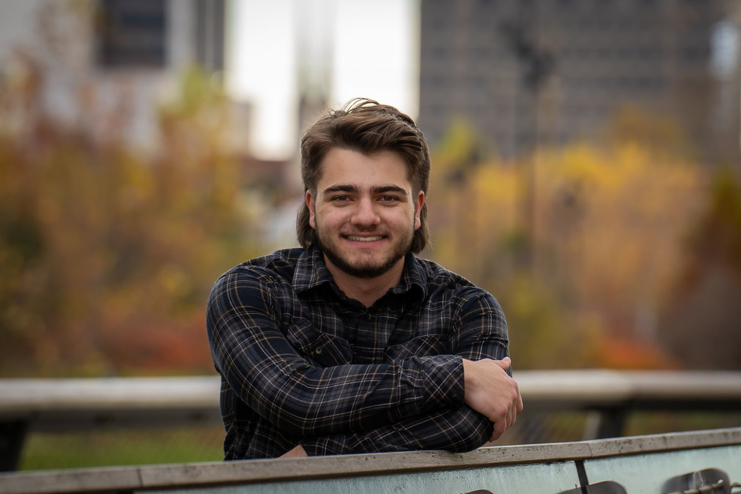 Smiling young man with beard leaning on railing with autumn cityscape in background.
