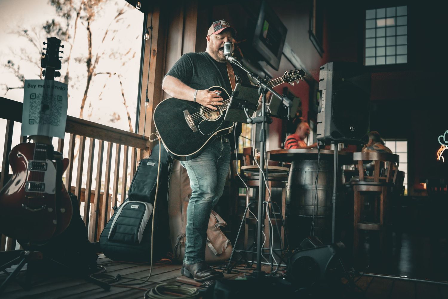 Bearded guitarist in black shirt plays acoustic guitar on rustic porch stage surrounded by vintage equipment.