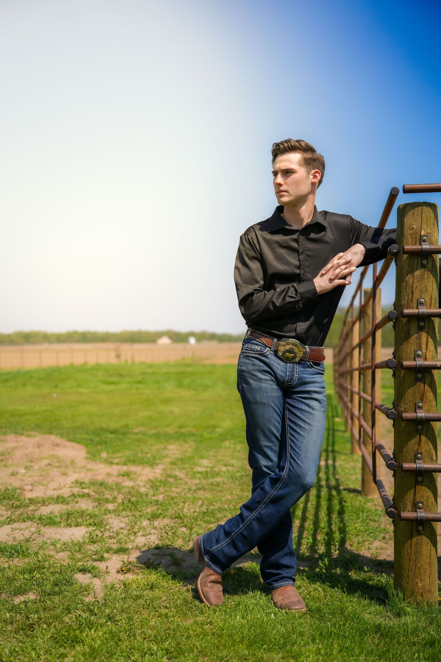 Young man in black shirt and jeans leans against a rustic farm gate in a green field.
